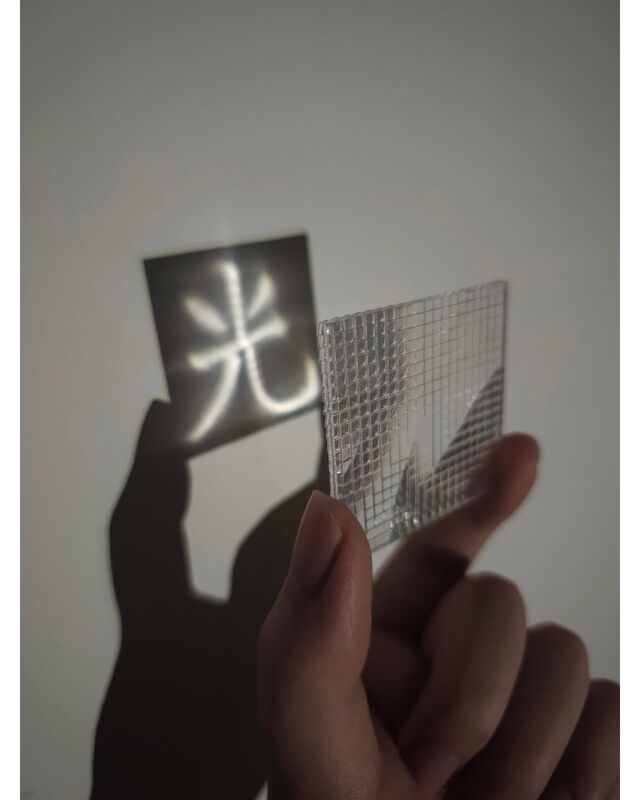 A faceted clear lens held up to a wall, projecting the character 光 in light inside a dark square